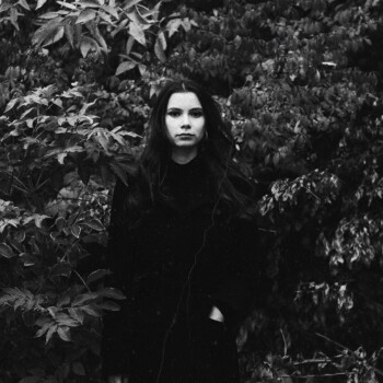 Photo by https://byabduls.com/work black and white photo of a girl wearing a black trench coat in front of leaves.