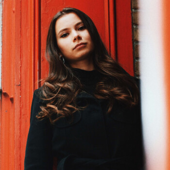 Photo by https://byabduls.com/work picture of a girl looking down at the camera in front of orange door frame wearing black trench coat